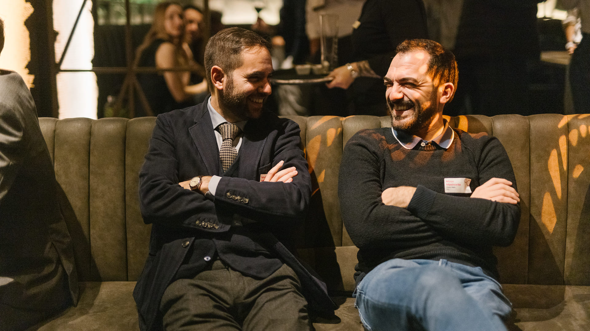 Two men laughing and talking on a couch at a social event.