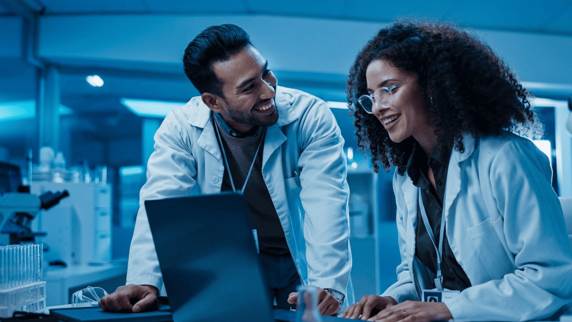 Zwei Wissenschaftler lächeln in einem Labor vor einem Laptop.